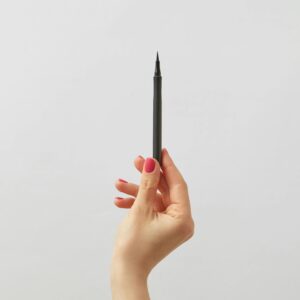 A hand with pink nail polish holding a black eyeliner pen against a white backdrop.